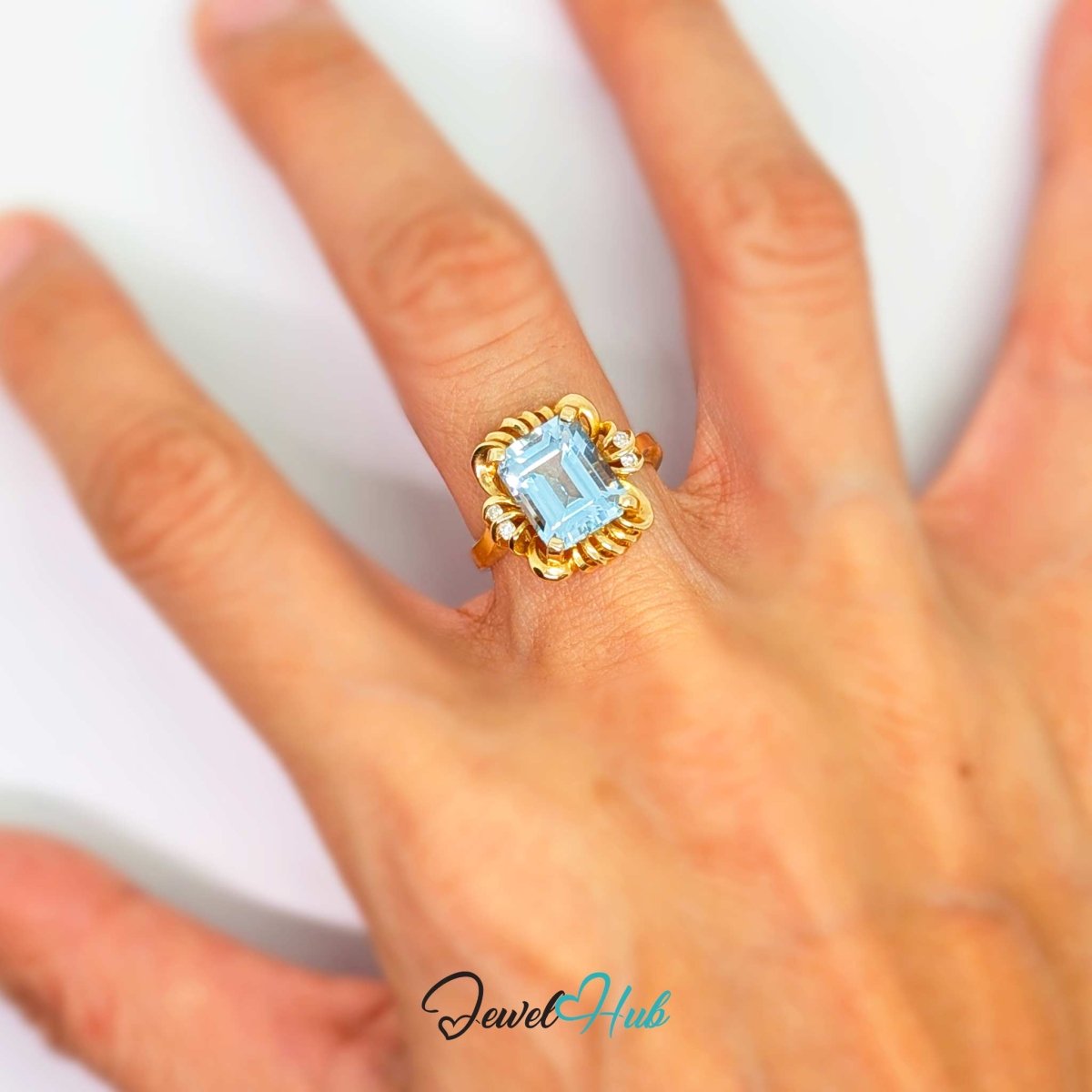 A 14k gold ring with a rectangular light blue gemstone and small clear accent stones, shown worn on a person's finger against a light background. The brand name 'JewelHub' is visible as a watermark at the bottom.