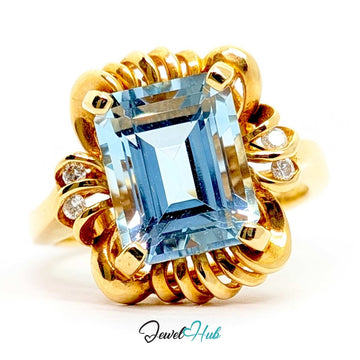 A 14k yellow gold ring featuring a large, cushion-cut light blue gemstone in a prong setting, surrounded by a textured, swirling gold halo with small accent diamonds on the sides, set against a plain white background.
