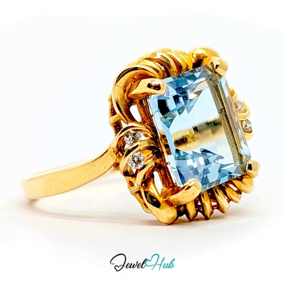 A 14k yellow gold ring featuring a large rectangular light blue gemstone with decorative scrollwork detailing and small accent stones, shown on a white background with a 'JewelHub' watermark.