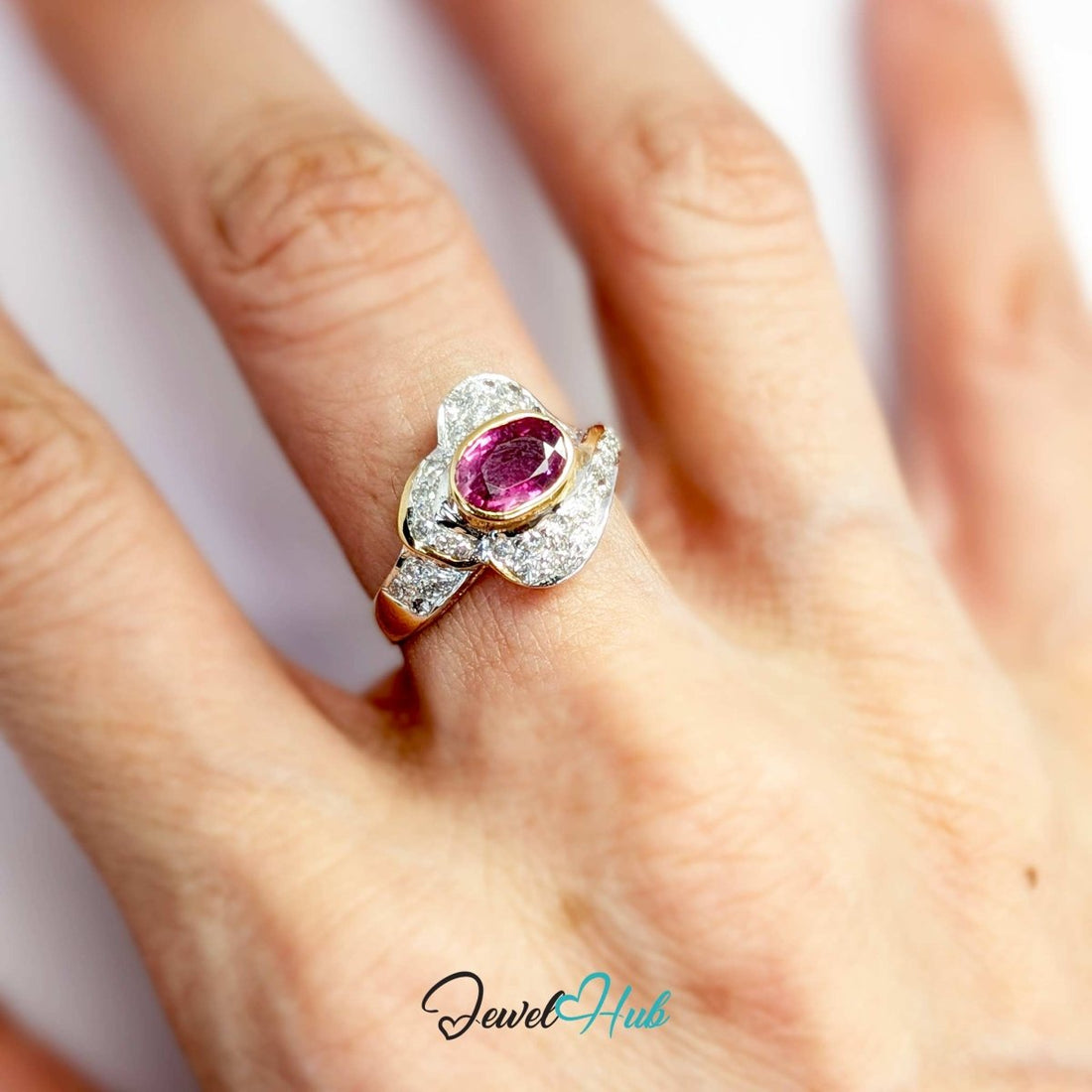 A solid 14k gold and white gold ring featuring a large oval-cut pink gemstone in the center, surrounded by pavé-set clear stones and accented with yellow gold details, shown on a white background with a 'JewelHub' watermark.