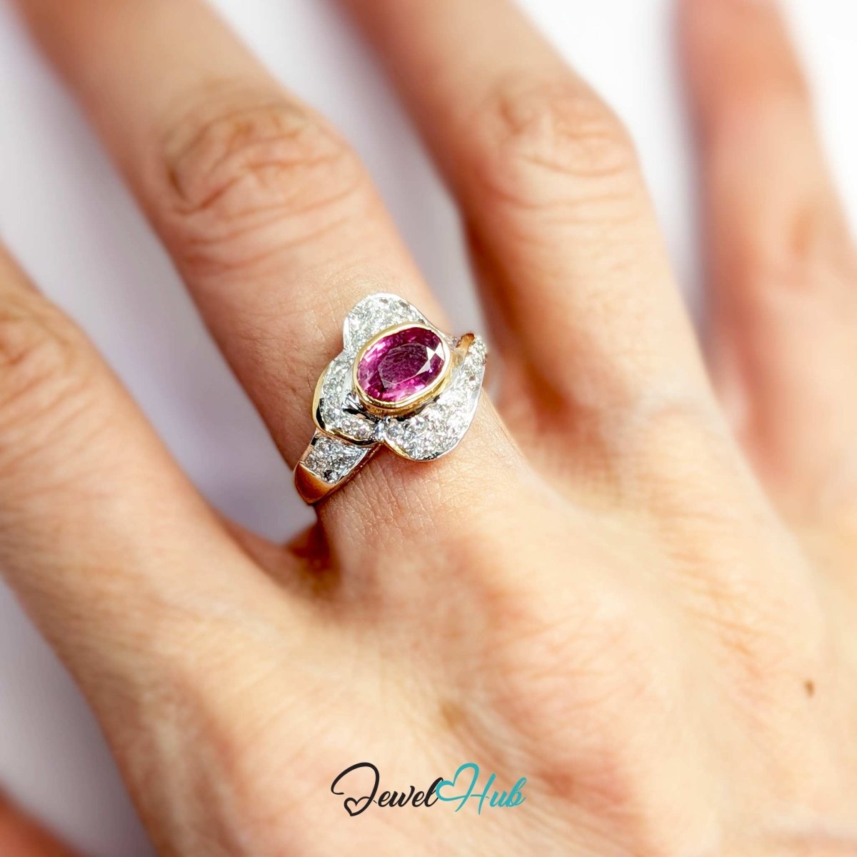 A close-up of a hand wearing a floral-shaped solid 14k ring with a central oval pink gemstone and surrounding clear stones in a silver-toned setting. The brand name 'JewelHub' is visible as a watermark at the bottom.