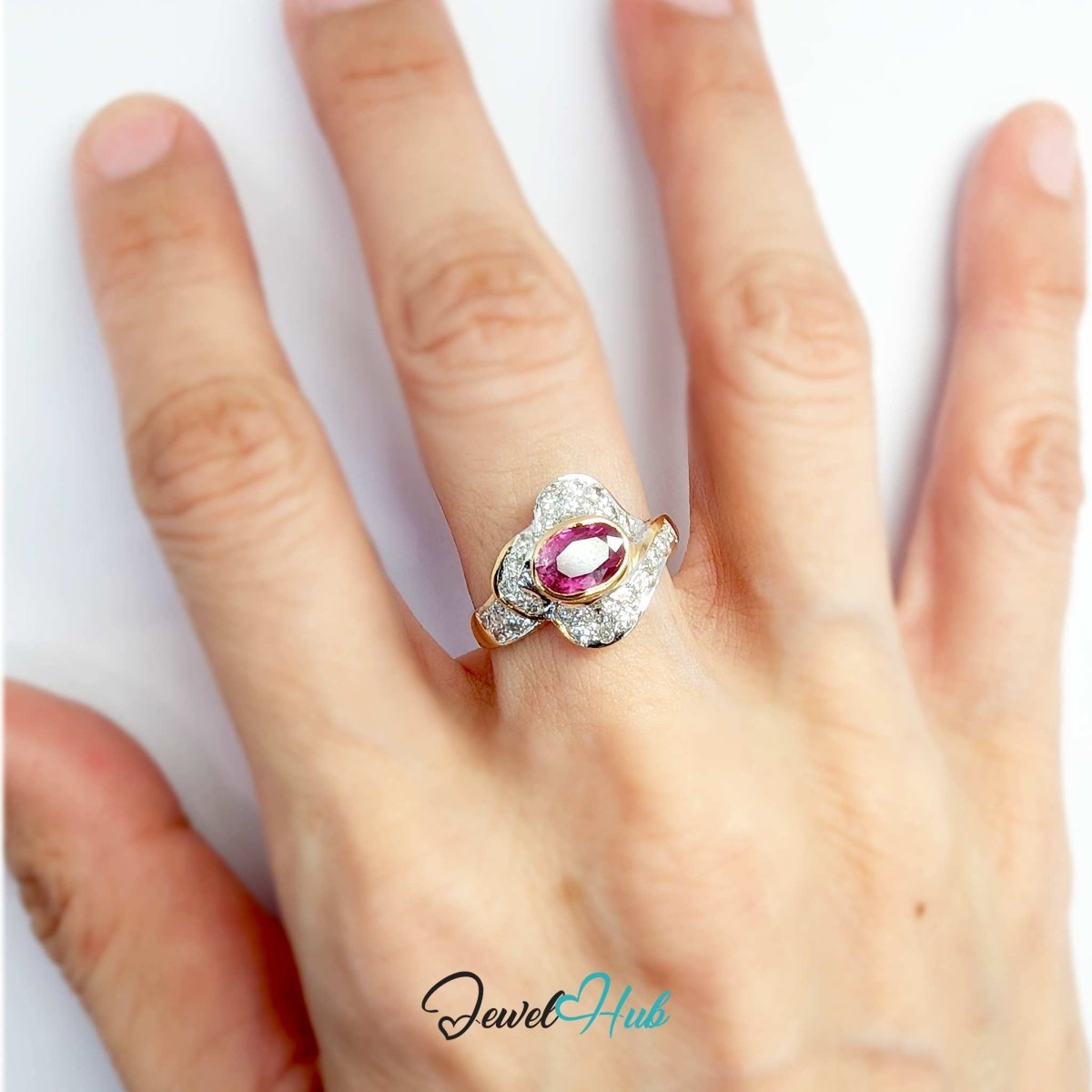 A solid 14k ring with a central oval pink gemstone and surrounding clear stones, shown worn on a hand against a white background. A 'JewelHub' watermark is visible at the bottom.