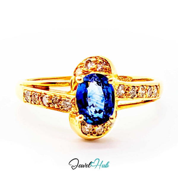 A yellow 14k gold ring featuring a large oval-cut blue sapphire center stone, surrounded by a halo of small clear stones, with additional clear stones set along the split shank. The ring is photographed on a clean white background with a 'JewelHub' watermark at the bottom.
