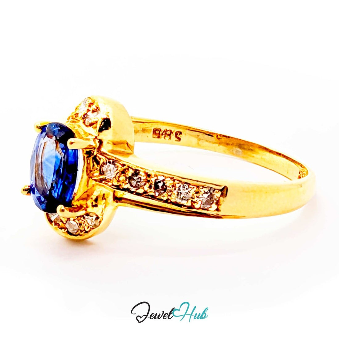 A yellow 14k gold ring featuring a large oval-cut blue sapphire center stone, surrounded by a halo of small clear stones, with additional clear stones set along the split shank. The ring is photographed on a clean white background with a 'JewelHub' watermark at the bottom.