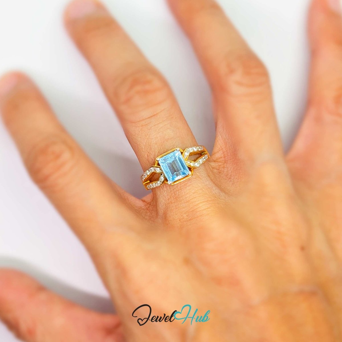 A 18k gold ring with a rectangular light blue gemstone and small clear accent stones, shown worn on a person's hand against a white background.