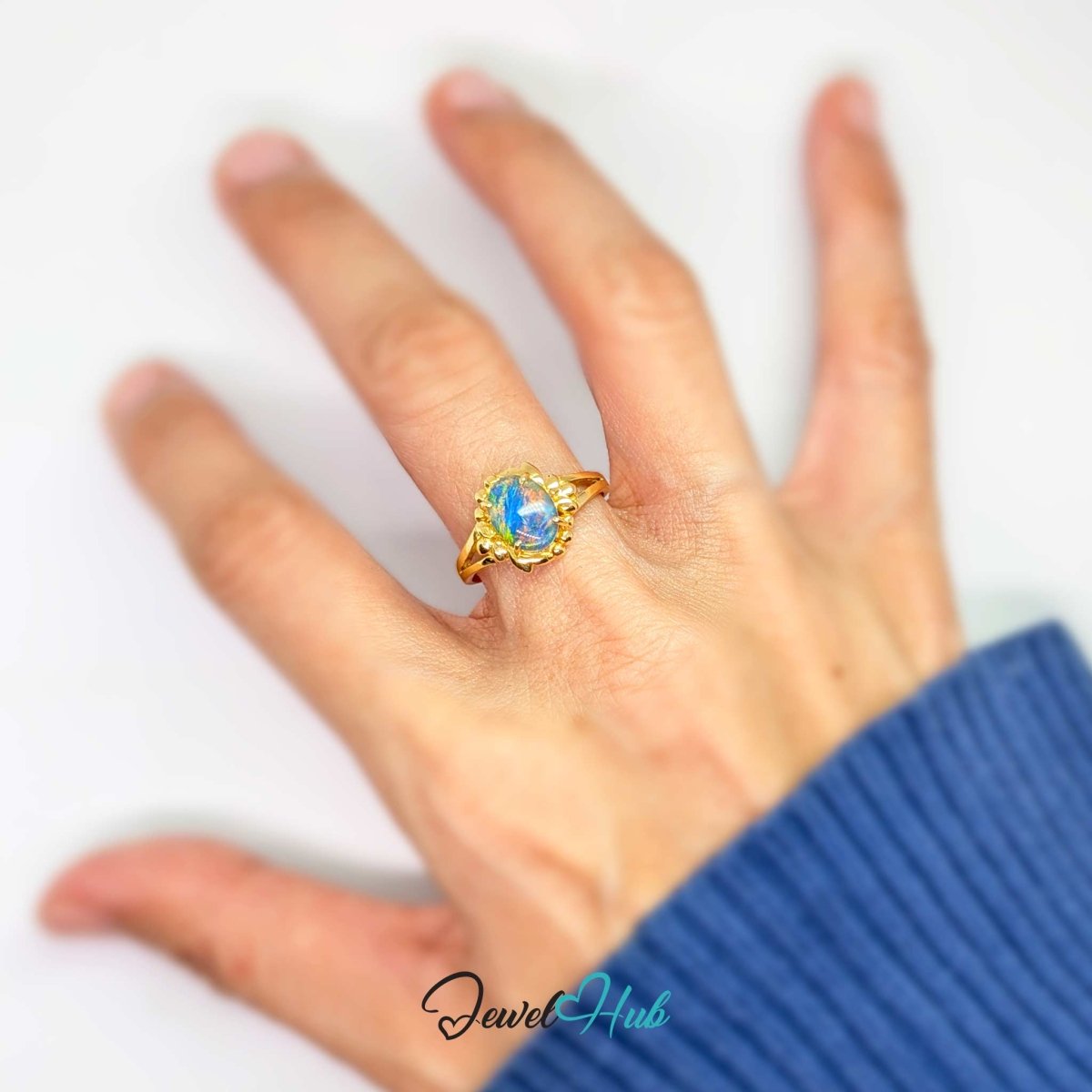 A 18k gold ring with a blue-green opal-like cabochon stone, worn on a person's ring finger. The hand is shown against a plain white background, wearing a blue ribbed sweater sleeve.