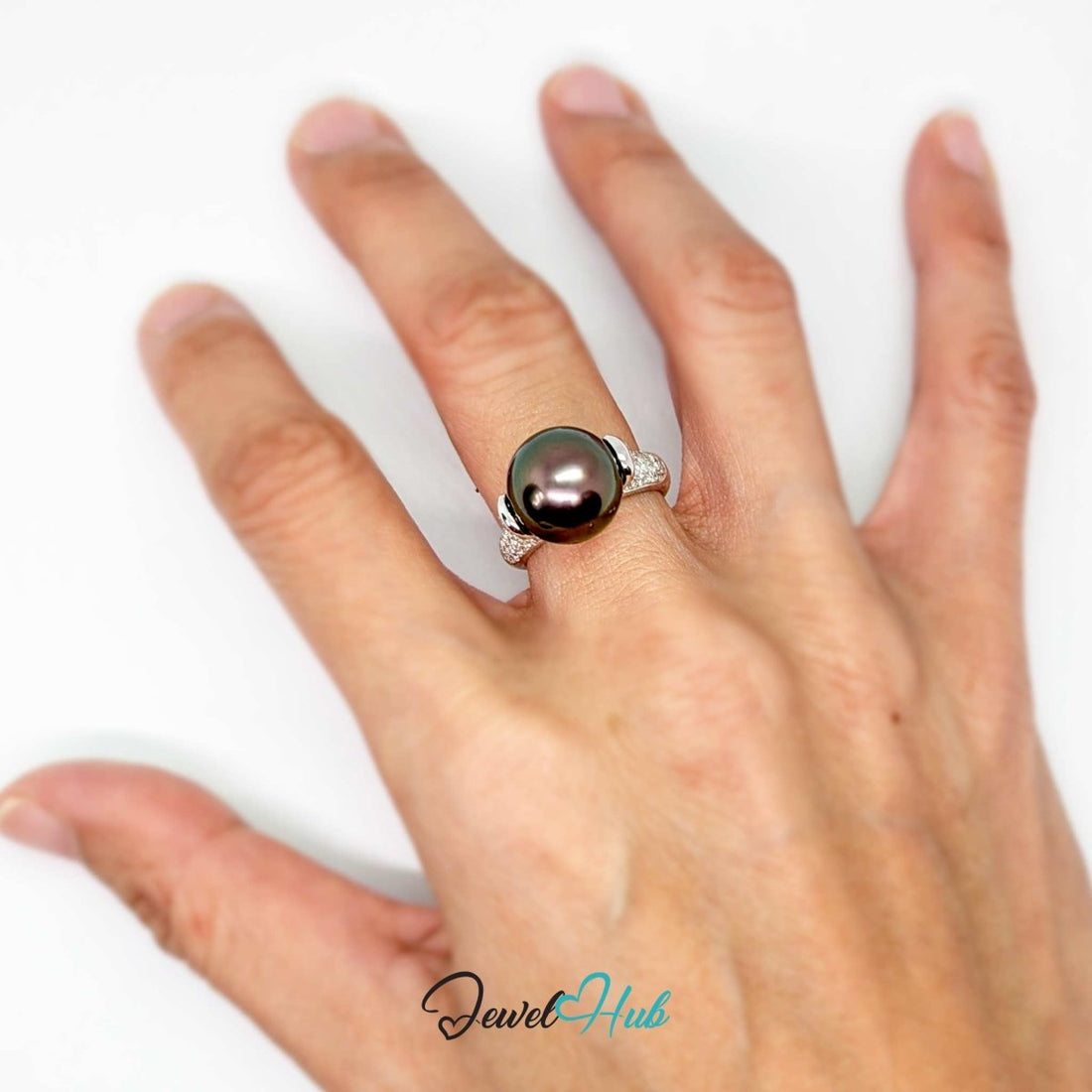 A 18k white gold featuring a large, round, dark green-black Tahitian pearl with iridescent pinkish-purple reflections, accented by pavé-set clear stones on the band, shown on a white background with a 'JewelHub' watermark.