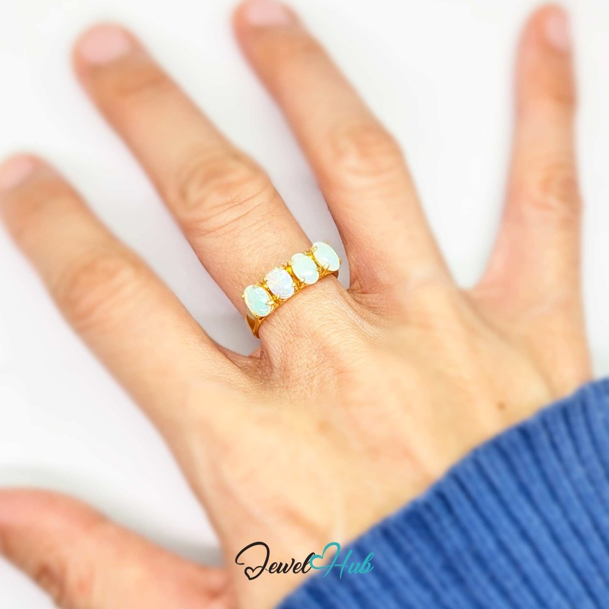 A gold 18k ring with four oval white opal gemstones worn on a person's ring finger, shown against a white background with a blue ribbed sweater cuff visible.