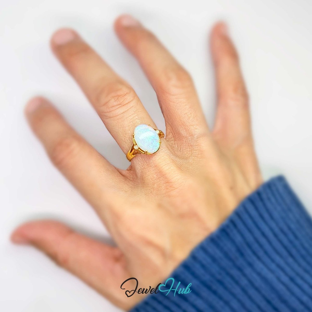 A 18k gold ring with an oval light blue opal gemstone worn on a person's ring finger, shown against a plain white background with a blue ribbed sweater cuff visible.