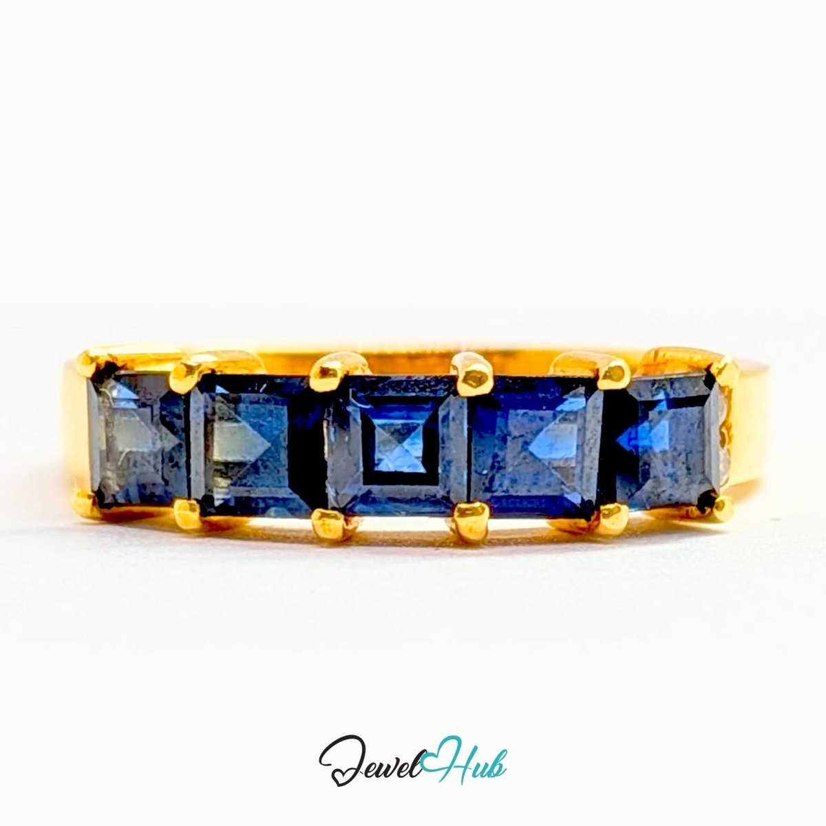 A yellow 18k gold ring featuring a row of five square-cut dark blue sapphires, set against a plain white background with a 'JewelHub' watermark at the bottom.