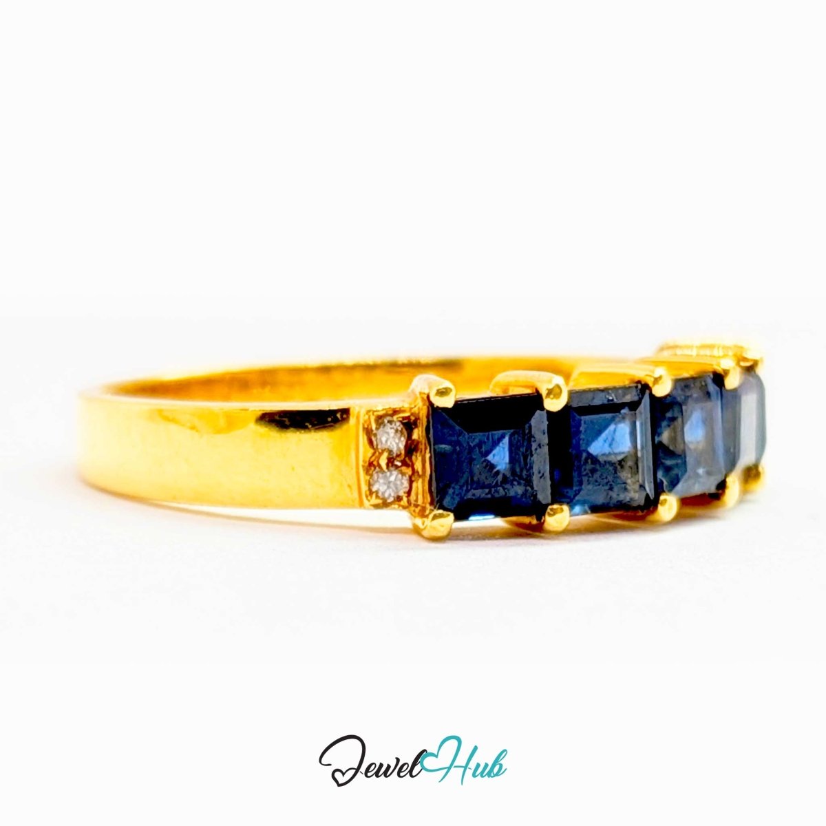 A yellow 18k gold ring featuring a row of square-cut dark blue sapphires, accented by small clear stones on the sides, set against a plain white background with a 'JewelHub' watermark at the bottom.