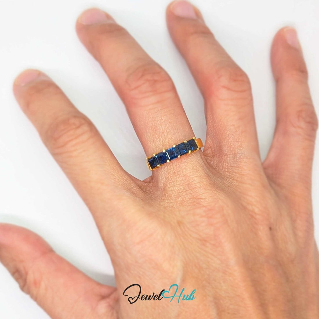 A yellow gold ring featuring five square-cut dark blue sapphires in a row, set against a plain white background with a 'JewelHub' watermark at the bottom.