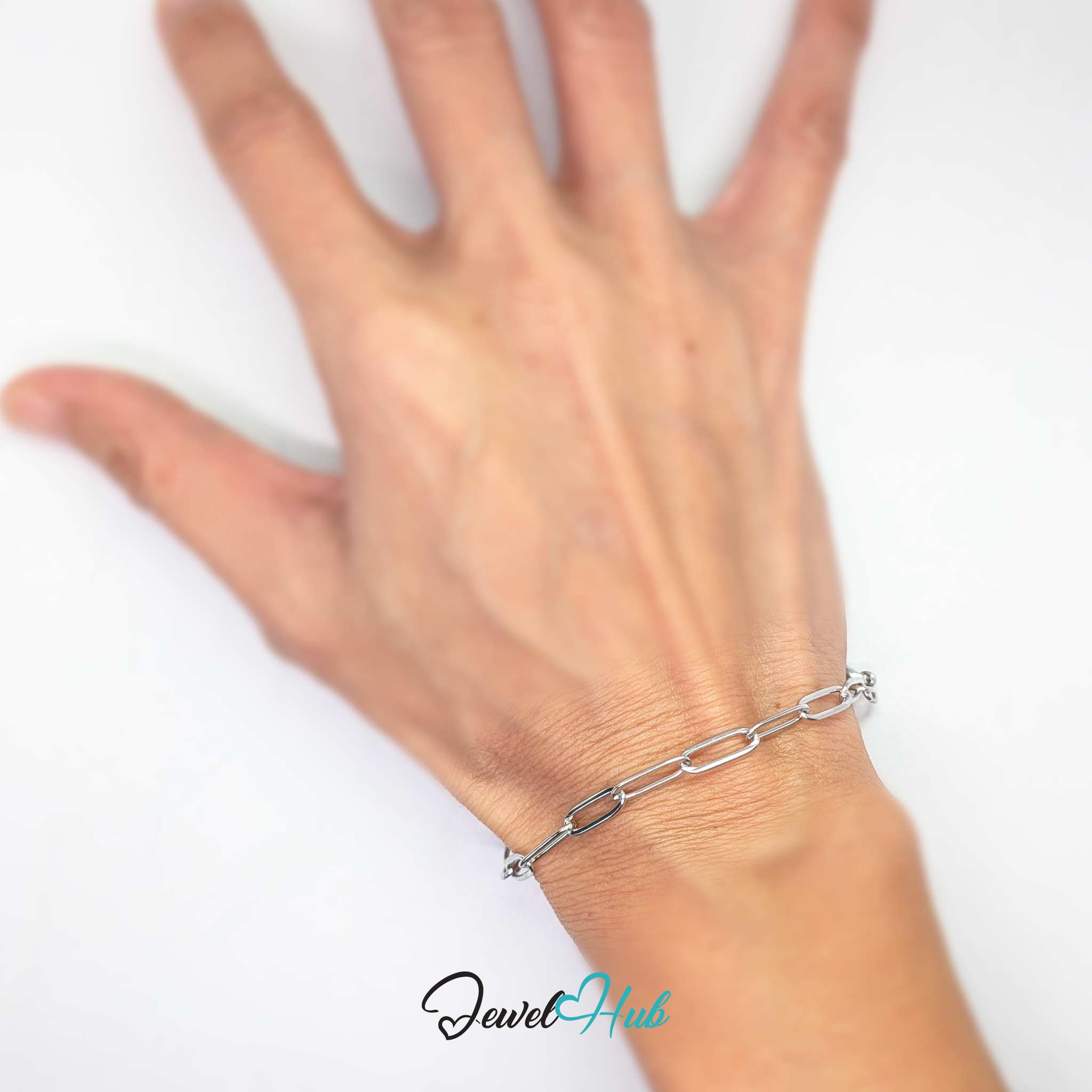 A close-up of a person's hand and wrist wearing a delicate silver chain bracelet with large paperclip-style links, set against a plain white background with a 'JewelHub' watermark at the bottom.