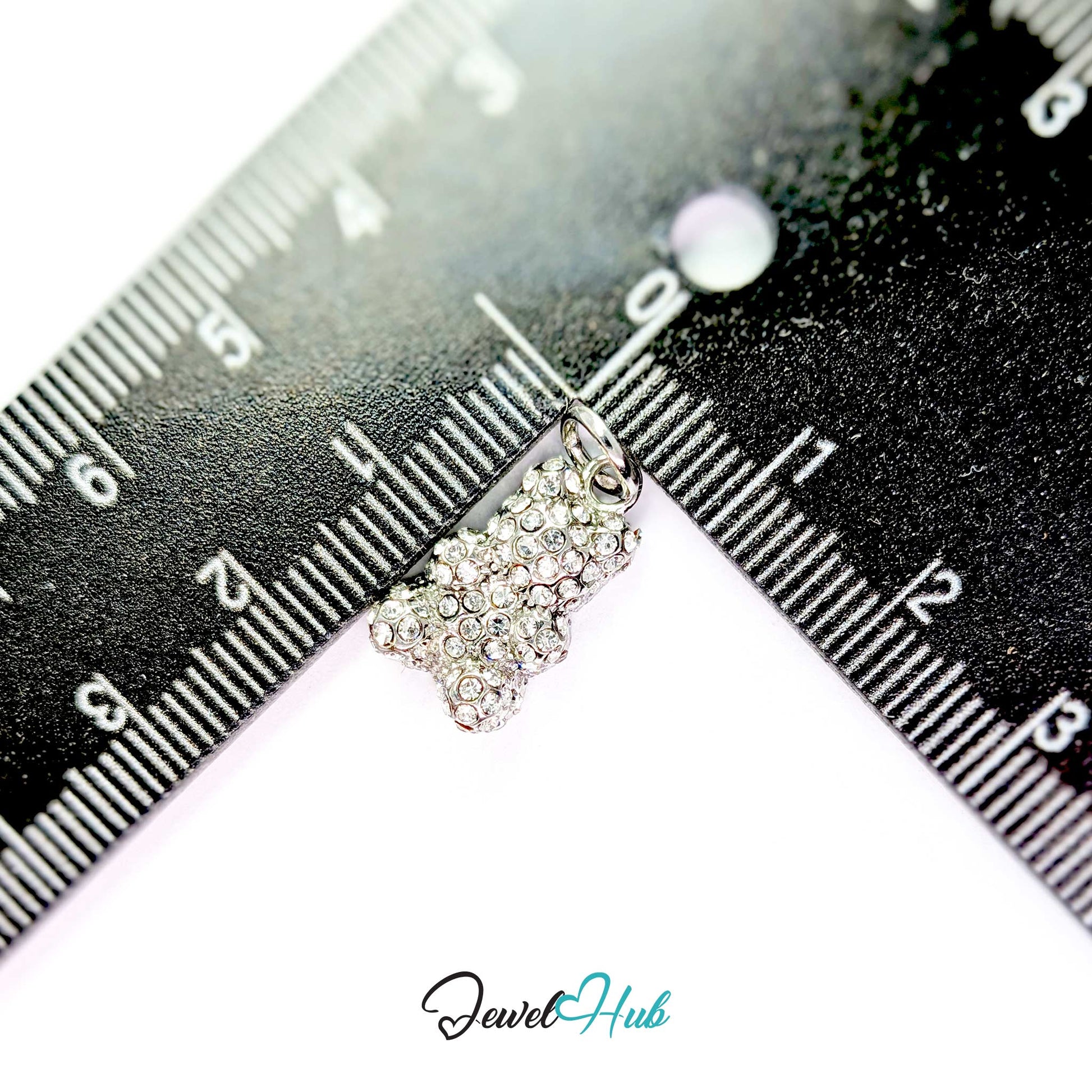 A small rhinestone-covered charm or pendant in a silver-tone setting, photographed close up between two black measuring rulers to show scale. The product is centered on a white background with a JewelHub watermark at the bottom.