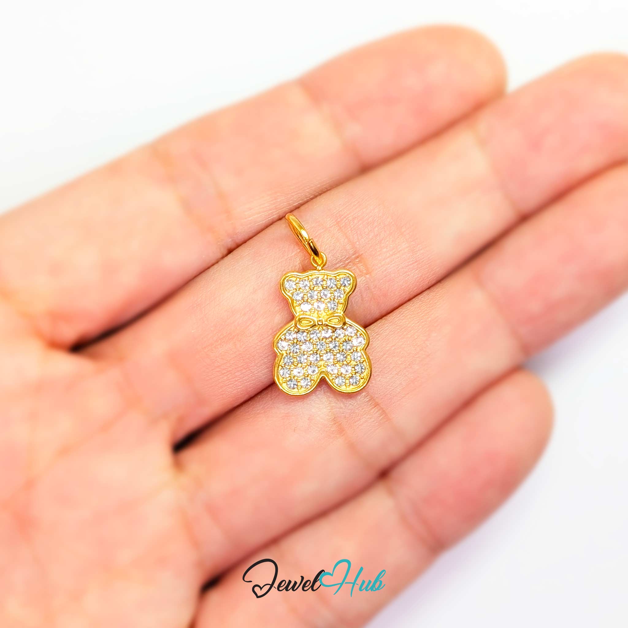 A small gold teddy bear pendant covered in clear pavé stones, held on an open hand for scale. The brand name 'JewelHub' is visible as a watermark at the bottom.
