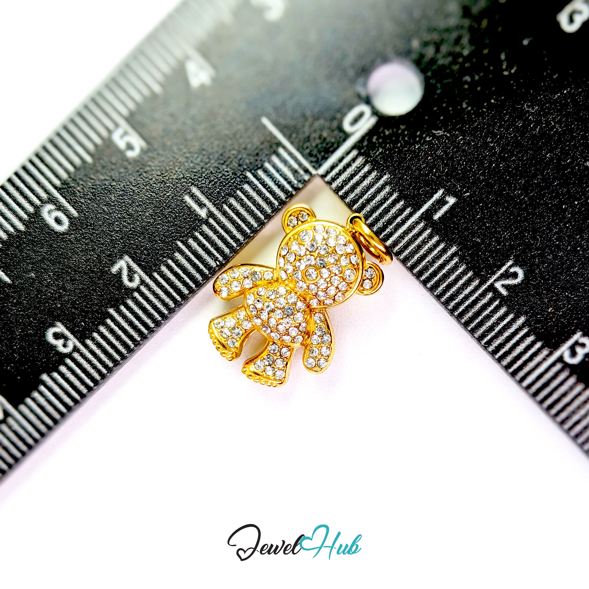 A small gold teddy bear pendant covered with clear rhinestones, shown between two black rulers for scale on a white background, with a JewelHub watermark at the bottom.