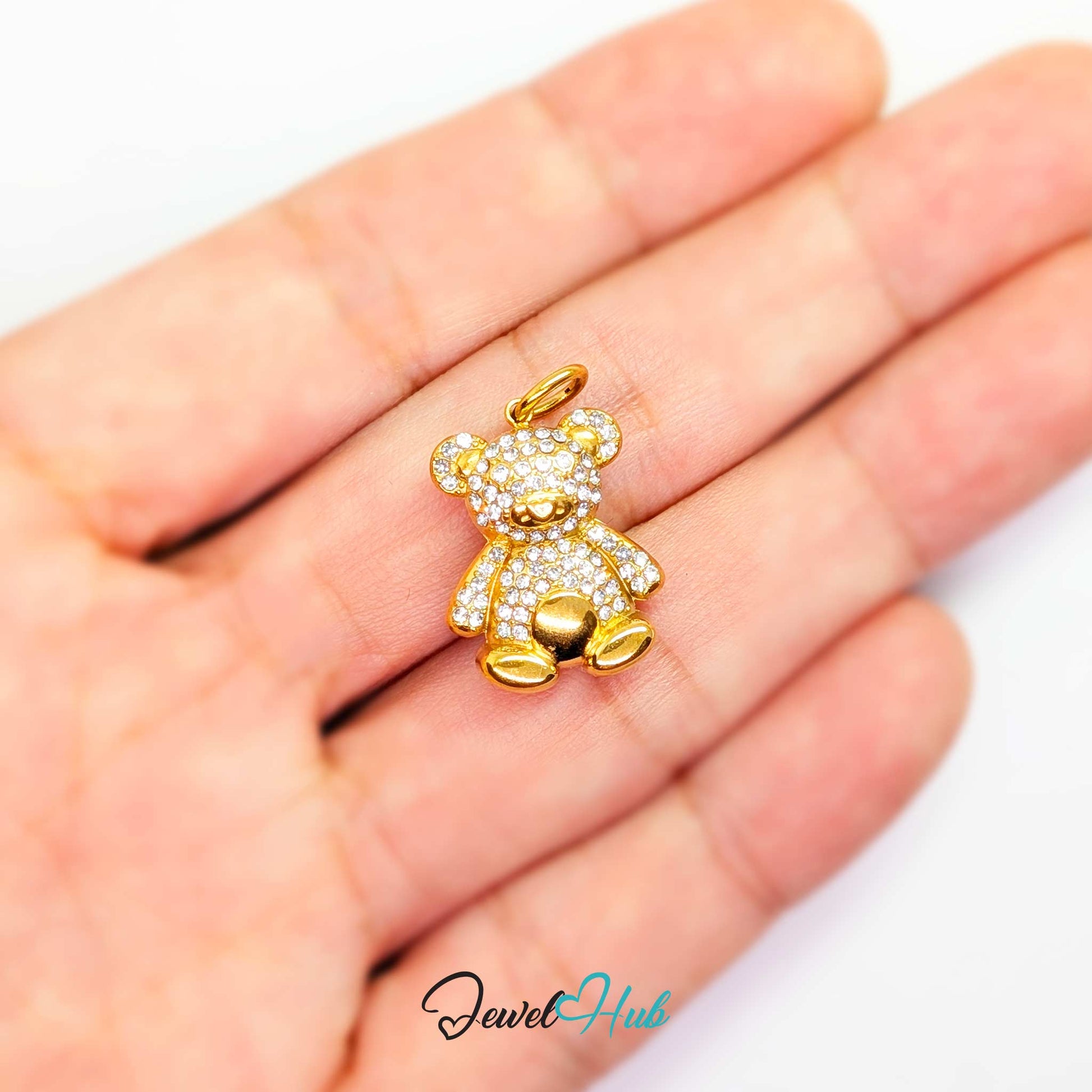 A small gold teddy bear charm paved with clear stones, resting on a person's palm to show scale. The brand name 'JewelHub' is visible as a watermark at the bottom.