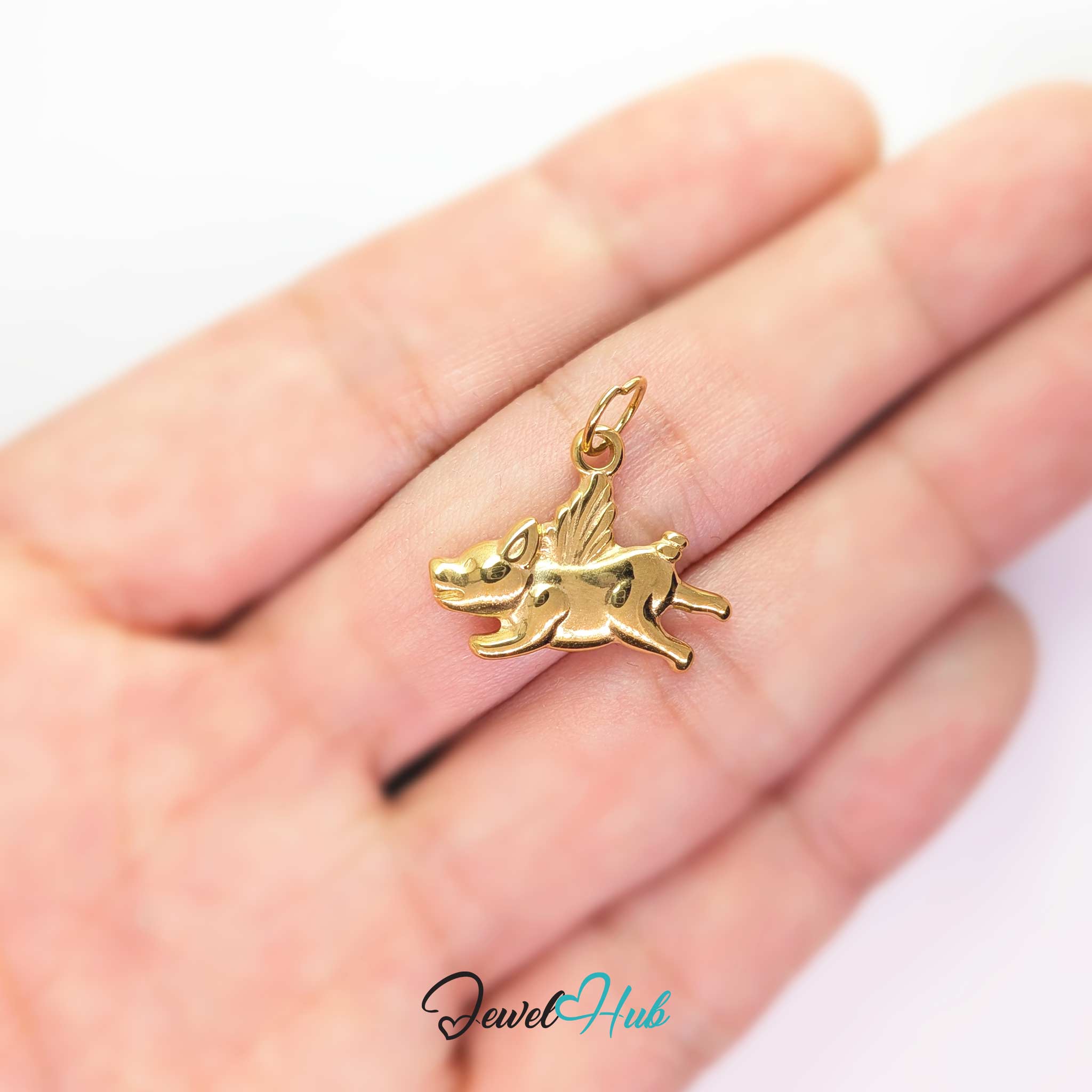 Gold flying pig charm held in the hand to show its size against a fingertip - JewelHub