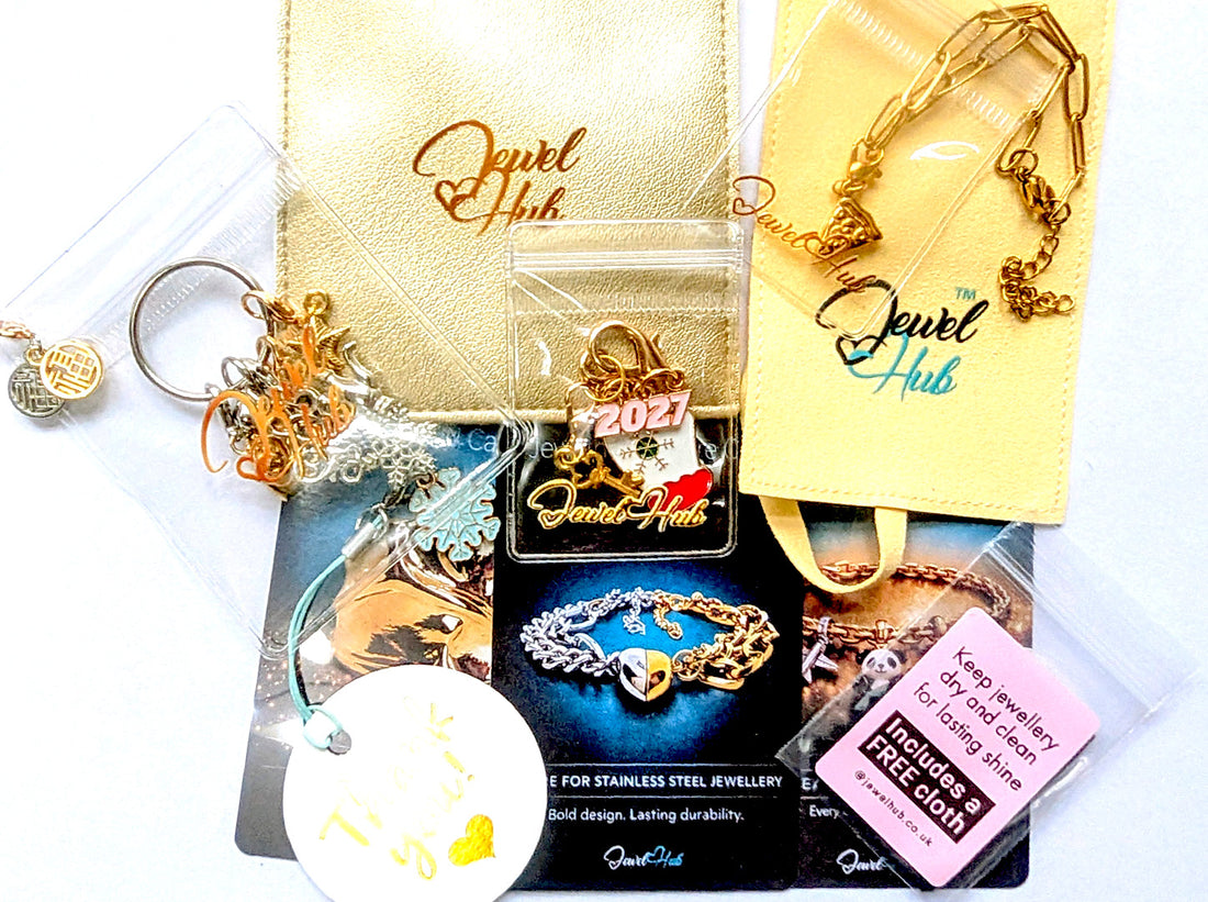 JewelHub Event Gift Bag contents laid flat: stainless steel paperclip chain bracelet, charm keyring with clip-on charms, gold and cream zip pouches, three material care cards, a Thank you tag and a free mini jewellery cloth
