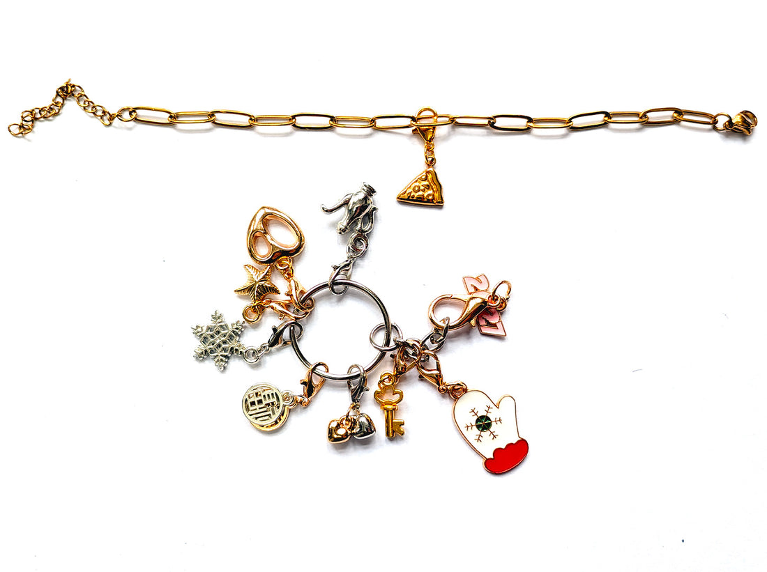 JewelHub Event Gift Bag contents laid flat: stainless steel paperclip chain bracelet, charm keyring with clip-on charms, gold and cream zip pouches, three material care cards, a Thank you tag and a free mini jewellery cloth