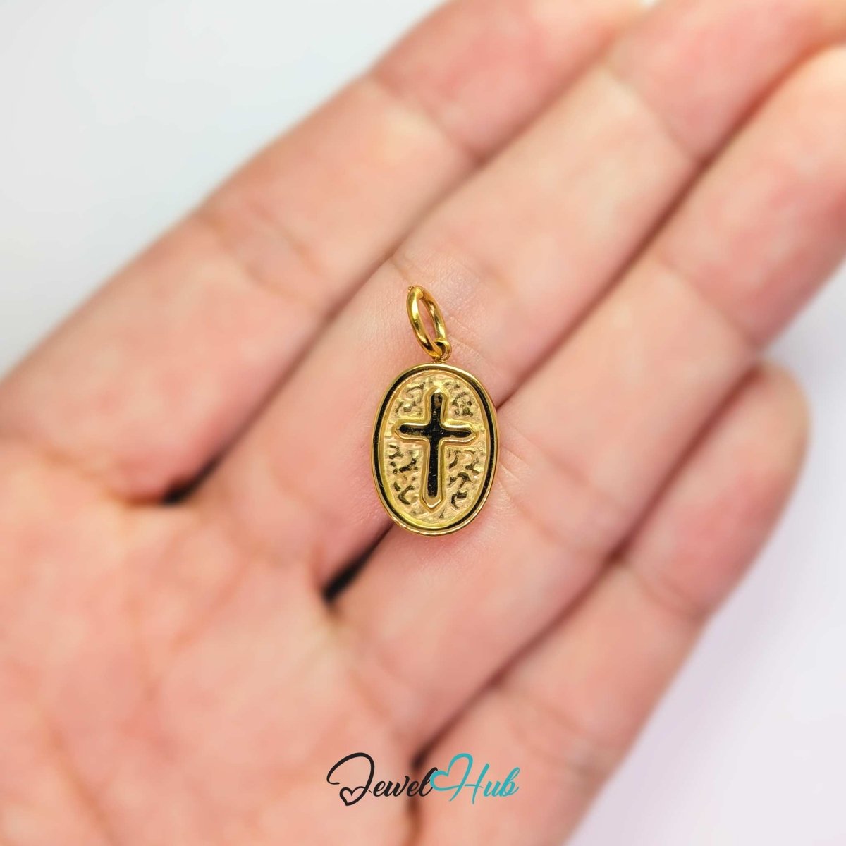 Oval cross medallion charm in gold stainless steel - held in the hand for scale - JewelHub