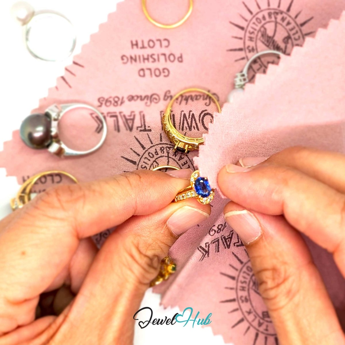 Hands buffing a gold sapphire and diamond ring with the pink Town Talk gold polishing cloth, other gold rings resting nearby.