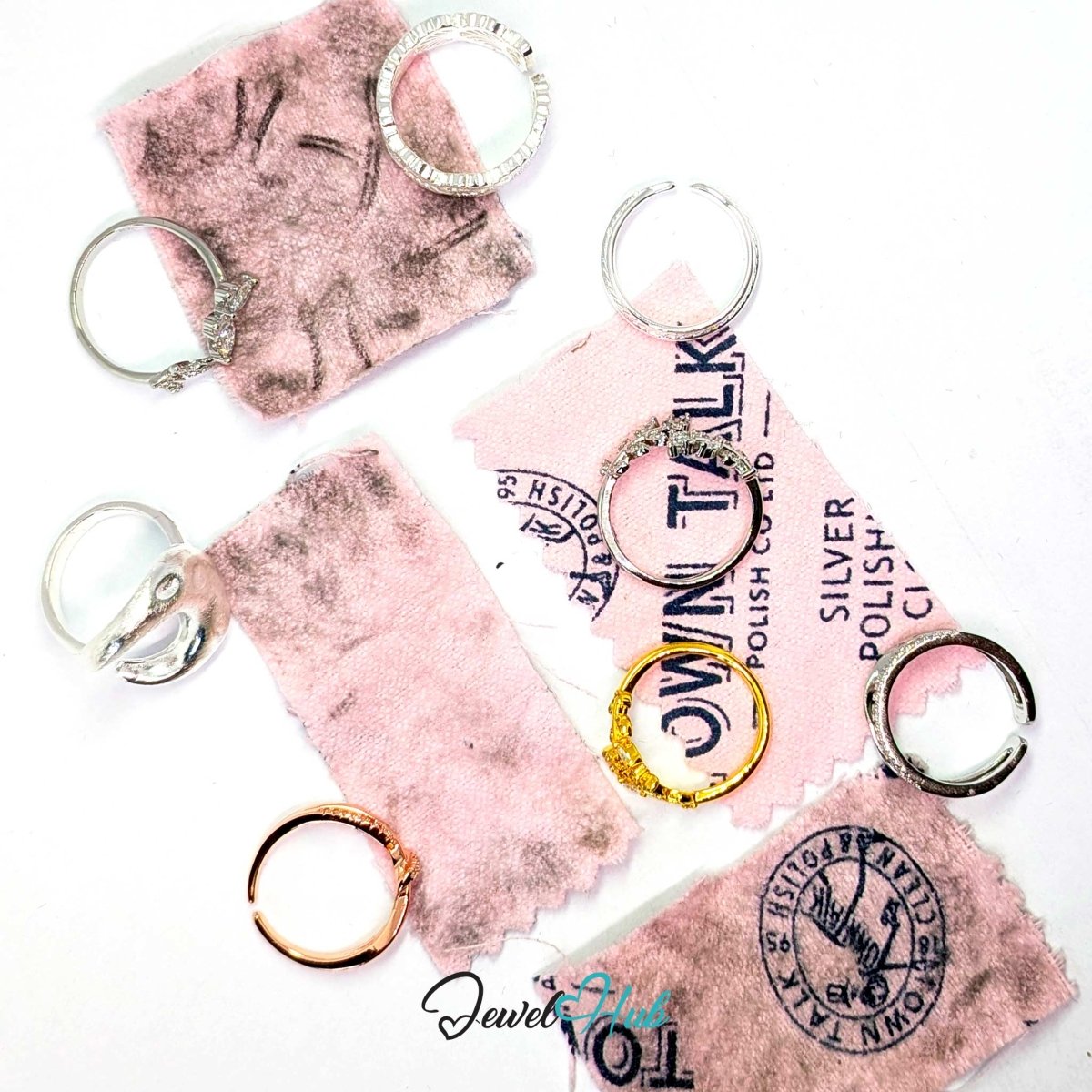 Several silver-toned metal key rings displayed on pink polishing cloths with visible tarnish and scuffs, arranged on a white background. Some rings are open C-style keychains, and one ring has a textured clear stone setting.
