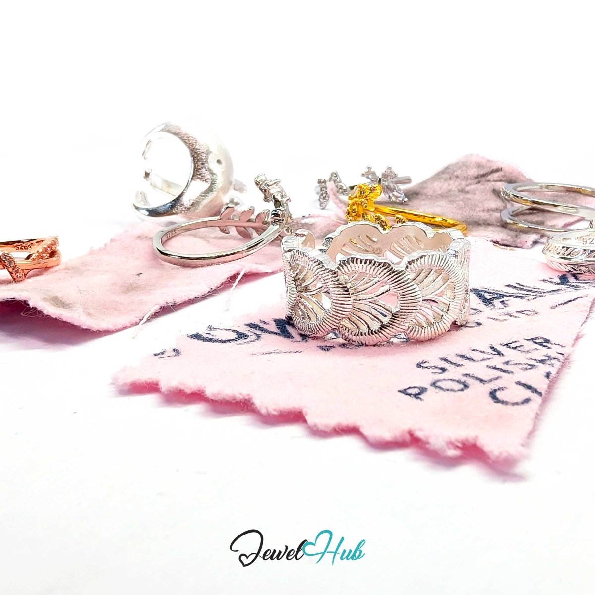 A silver ring with an intricate leaf or feather filigree design, displayed on a pink polishing cloth alongside other jewelry pieces on a white background. The 'JewelHub' logo is visible at the bottom.