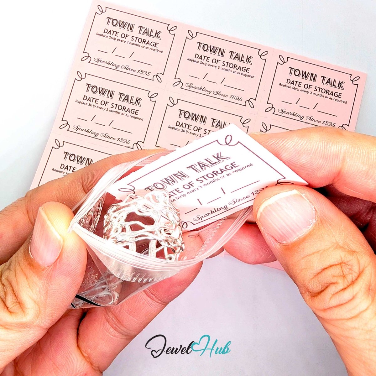 A close-up of hands holding a clear plastic bag containing a silver wire-wrapped pearl, with a pink 'Town Talk Date of Storage' label attached. More matching pink labels are stacked in the background on a white surface.