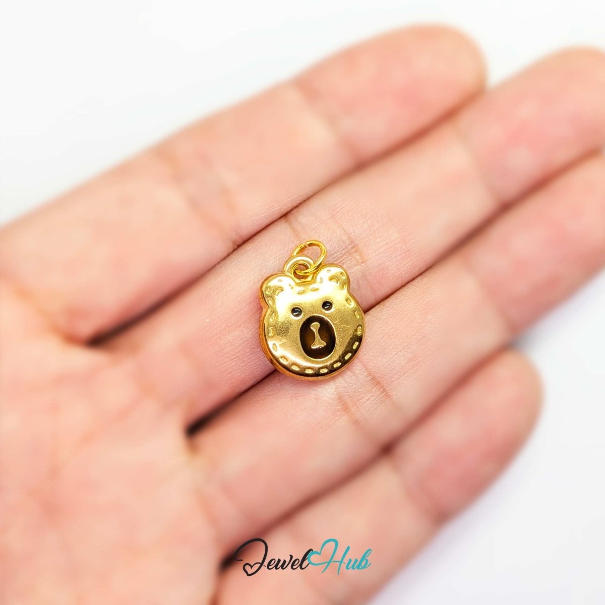 Gold Stitched Bear Head charm held in the hand to show its size against a fingertip - JewelHub